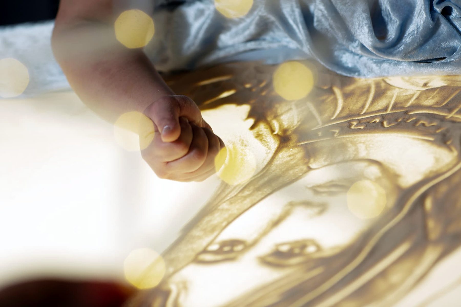 Hand zeichnet ein Gesicht in Sand auf dem Leuchttisch, im Vordergrund Lichtpunkte